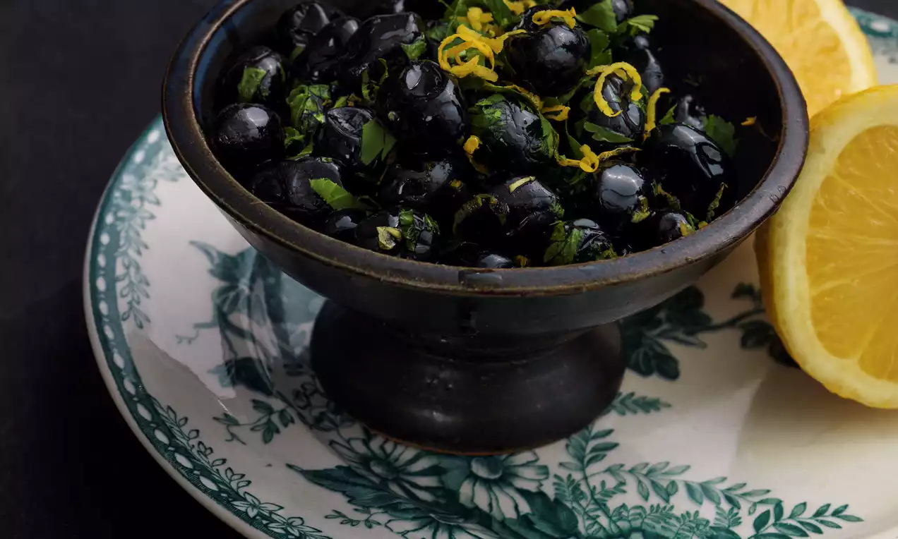 Svarta oliver i skål med citronzest och örter på dekorativ tallrik, bredvid halverad citron på mörk bakgrund