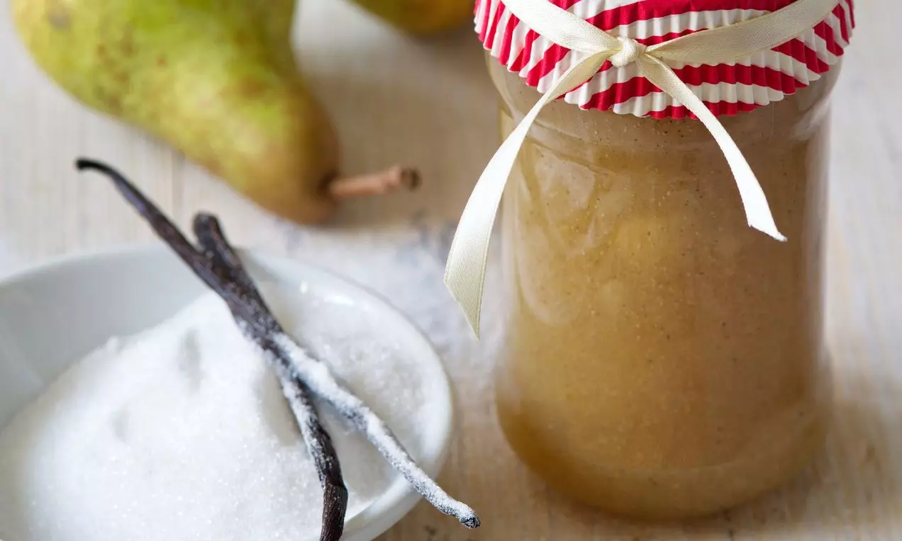 Hladké hruškové pyré jemne hnedej farby v sklenenej nádobe s červeno-bielym vrchnákom, vedľa vanilkových strukov, cukru a čerstvých hrušiek