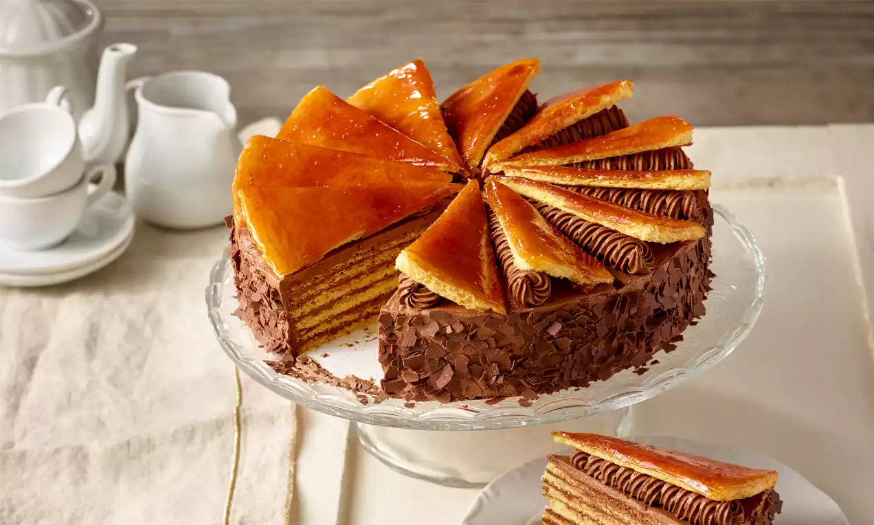 Mehrschichtige Dobostorte mit karamellglasierten Dreiecken, Schokoladencreme und Schokoraspeln auf Glasplatte, daneben Teller mit Kuchenstück