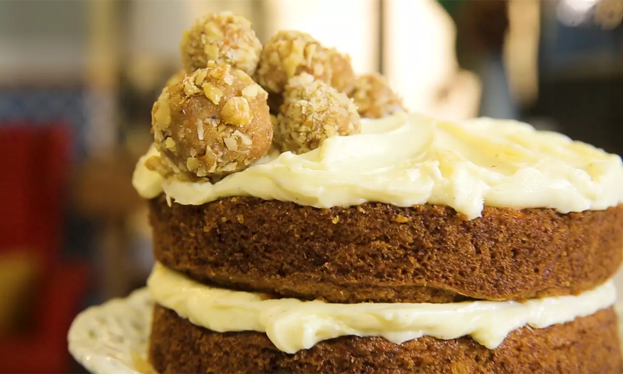 Двуслойна торта с наситено кафяв цвят, кремообразна глазура между пластовете и отгоре, украсена с ядкови топчета върху бяла поставка