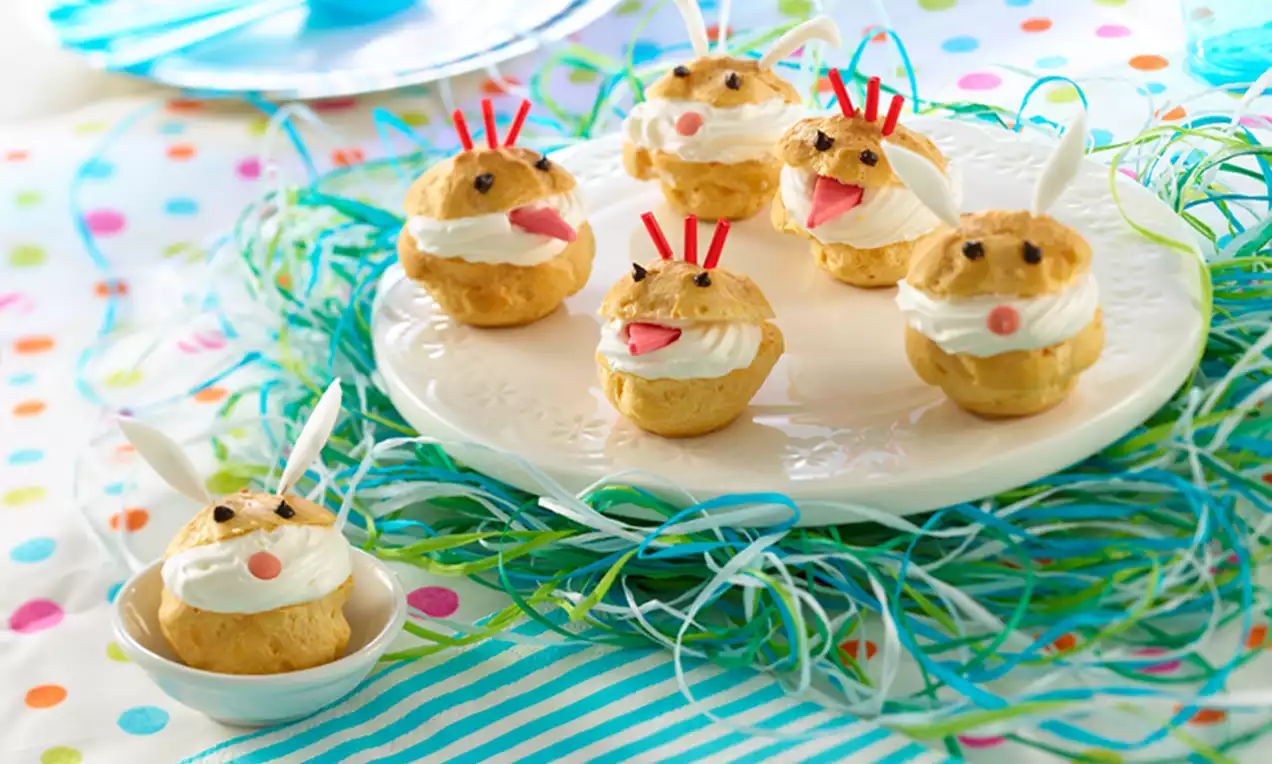 Profiteroles dorées remplies de crème blanche en forme de lapins, avec oreilles en sucre, yeux en chocolat et museaux roses, disposées sur une assiette blanche entourée de brins en papier coloré