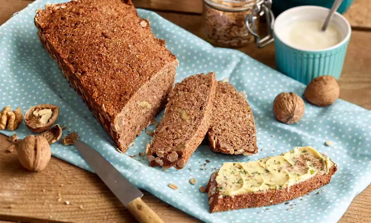 Rustikales Joghurt-Nussbrot mit brauner Kruste, angeschnitten auf einem blauen Tuch mit Butter bestrichen, Walnüssen und Messer drapiert