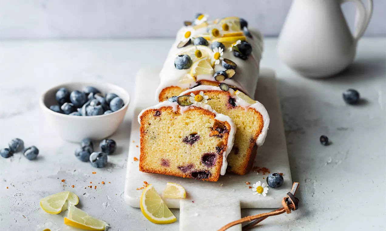 Picture - Saftiger Rührkuchen mit Blaubeeren und Zitronenscheiben, glasiert mit weißem Zuckerguss, dekoriert mit Kamillenblüten auf heller Holzplatte