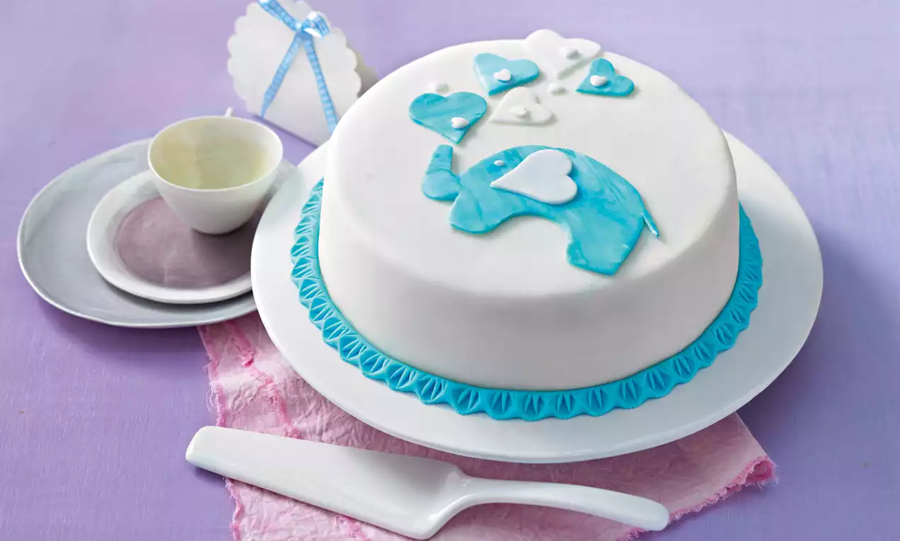 Runde Baby-Torte mit weißem Fondant überzogen, blauem Randdekor, Elefanten- und Herzmotiven, auf weißer Platte mit zartrosa Unterlage
