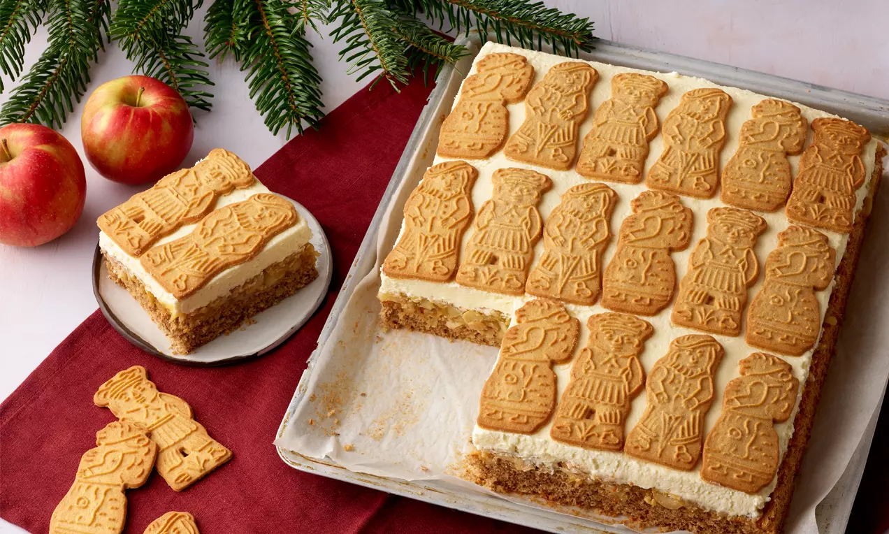 Ein rechteckiger Spekulatiuskuchen mit hellbraunem Boden, heller Creme und dekorativen Spekulatius obenauf auf rotem Tischläufer mit Äpfeln und Tannenzweigen