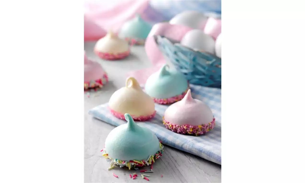 Pastel-toned meringues with swirl peaks, edges dipped in rainbow sprinkles, arranged on a light surface with a blue basket in the background