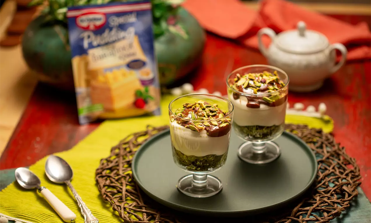 Десертът е в стъклени чаши с тъмен шоколадов крем, слой кремообразна сметана, зелена основа и поръска от ситно нарязани ядки