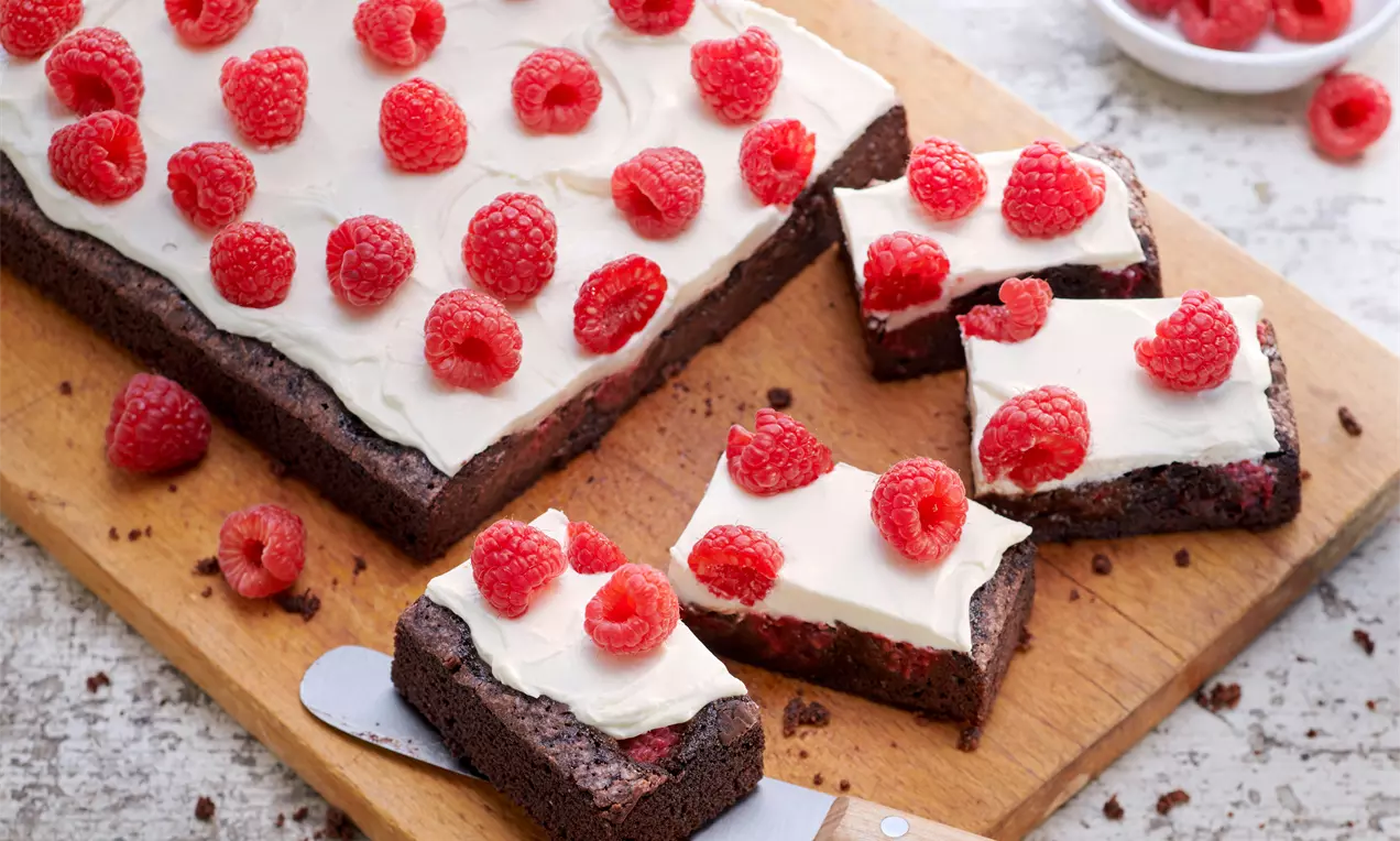 Saftiger, dunkelbrauner Brownie mit glatter, weißer Creme und leuchtend roten Himbeeren, auf einem Holzbrett angerichtet