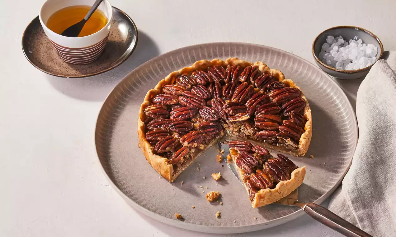 Kleiner Pecan-Pie mit goldbraunem Rand und glänzenden Pecannüssen in runder Form, angeschnitten auf einem hellen Teller mit Tee und Salzschale
