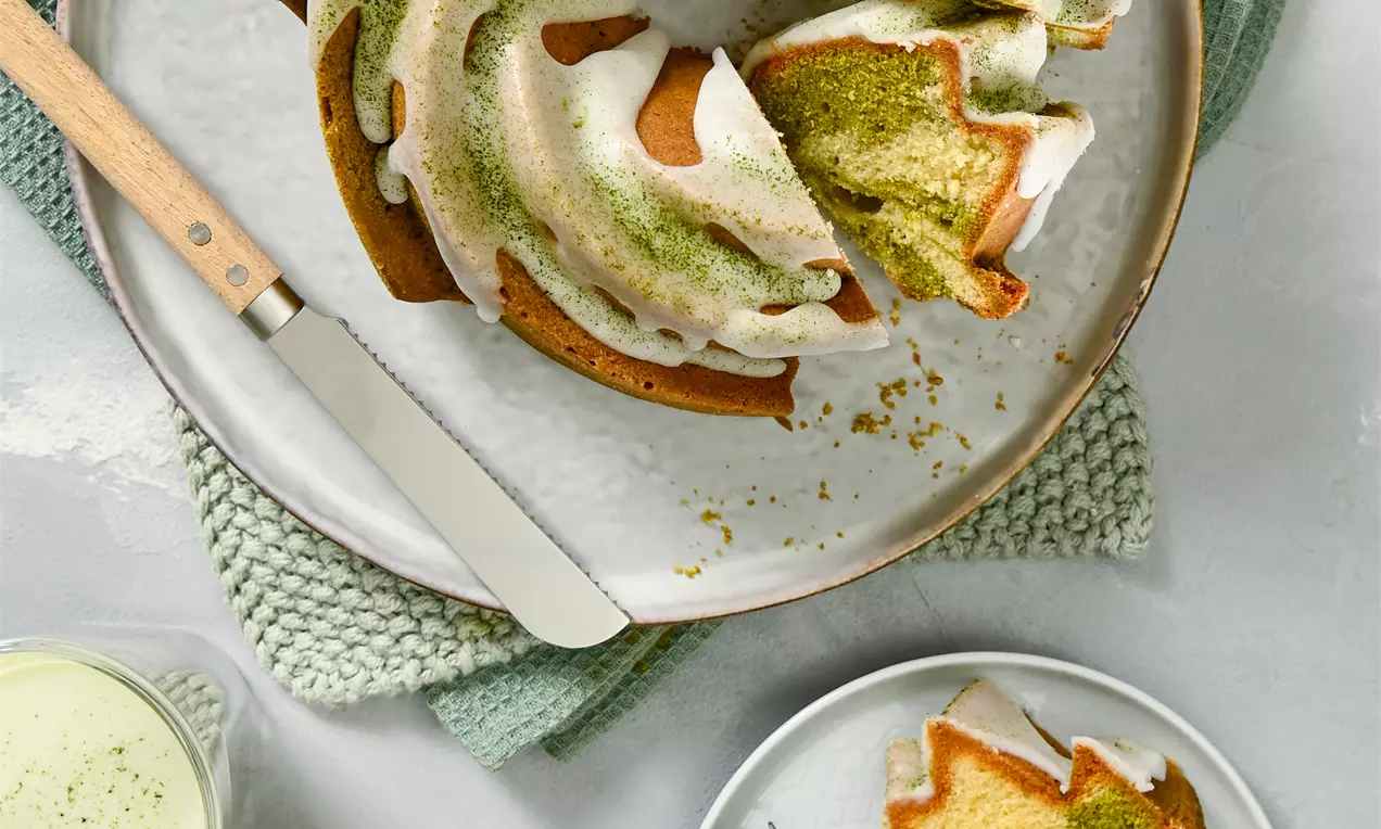 Bundt kolač spiralnog oblika s glazurom od bijele čokolade i posipom od zelenog matcha praha na tanjiru, uz servirane kriške