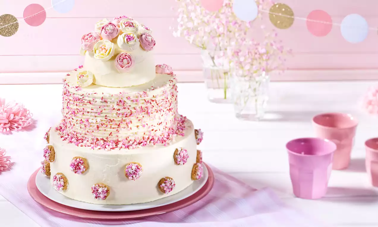 Mehrstöckige Torte mit heller Creme, rosa und goldenen Zuckerdekor, Blüten aus buntem Fondant und kleinen Keksen auf rosafarbenem Teller