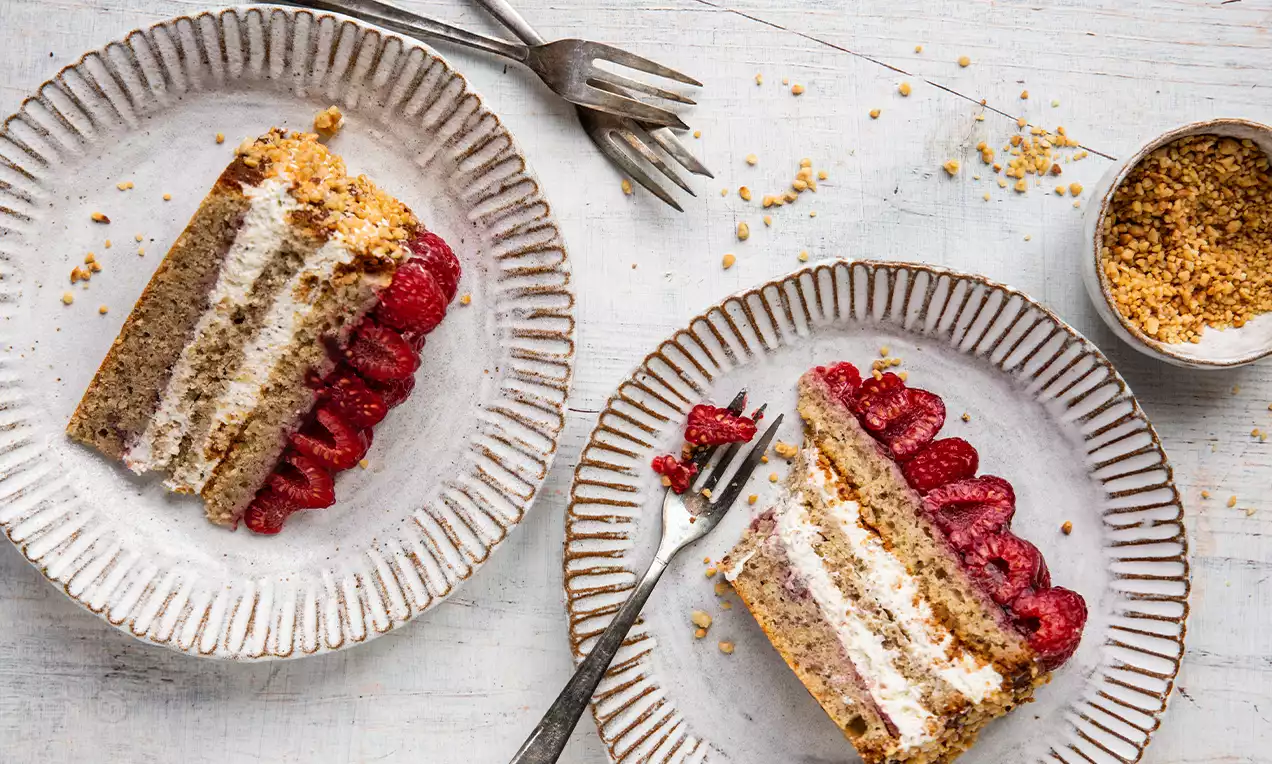 Tranches de gâteau aux noix moelleux avec une crème légère, garnies de framboises rouges vives et de miettes dorées sur des assiettes côtelées