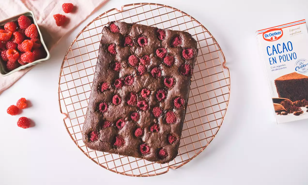 Brownie rectangular de intenso color marrón con textura húmeda cubierto de frambuesas frescas distribuidas sobre una rejilla dorada
