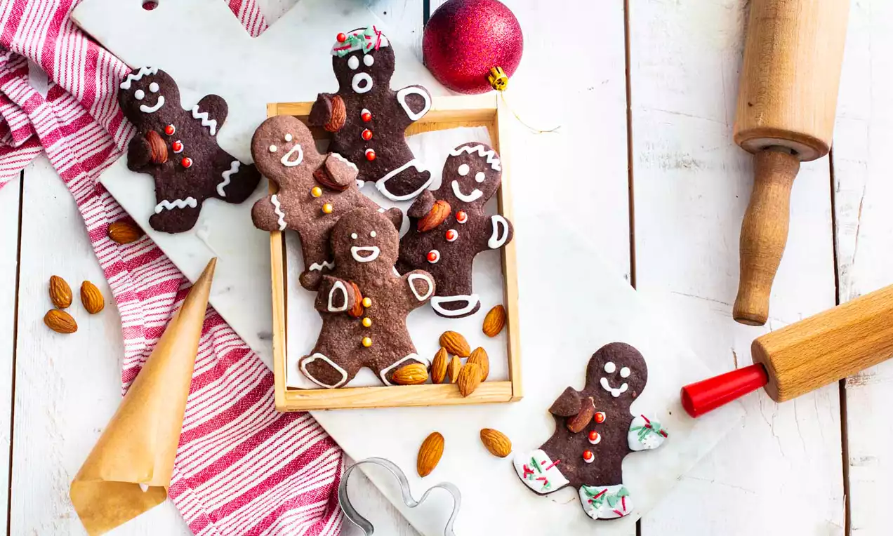 Honningkagemenn-småkager med mørkebrun overflate, hvit glasur og farvede detaljer ligger i en træbakke omgivet af mandler og julepynt