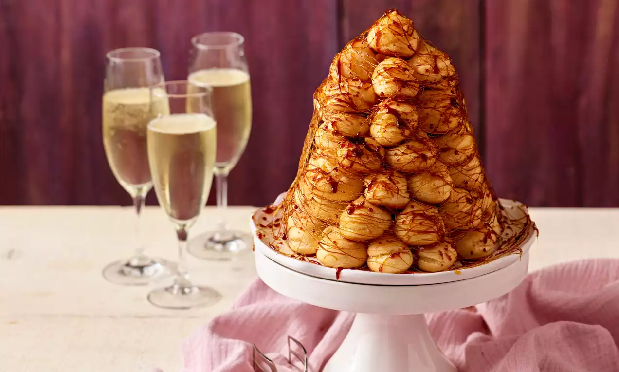 Zlatorjava piramida nežno hrustljavih profiterolov, prepletena s tankimi nitmi karamele na belem podstavku s svetlečo površino