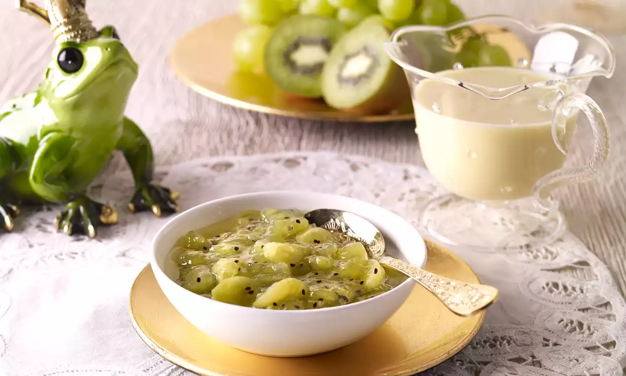 Kiwi-Weintrauben-Grütze mit grüner, glänzender Textur in einer weißen Schale auf einem goldenen Teller mit Sahnekanne und Dekoration