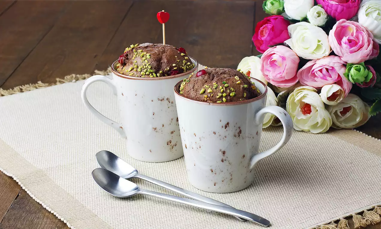 Ahşap masada açık renk hasır örtü üzerinde yan yana duran iki beyaz kupada çikolatalı kek, üstü fıstık ve kırmızı süslerle kaplı