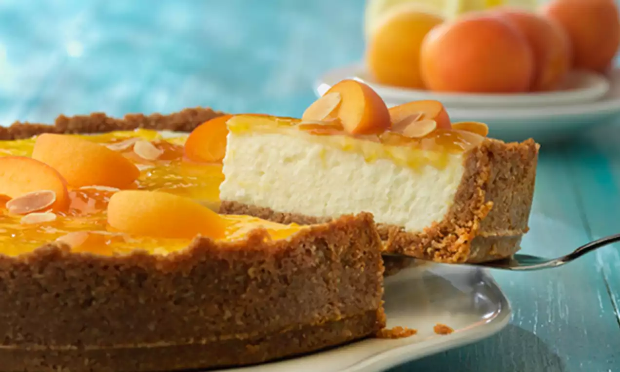 Tarta de queso cremosa sobre base dorada de galleta con cubierta brillante de albaricoque, gajos frescos y láminas de almendra en un plato