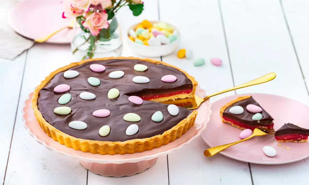 Шоколадов тарт с гладка тъмна глазура и ярки малини върху кадифен червен слой, поднесен в светлорозова чиния с цветни декоративни бонбони