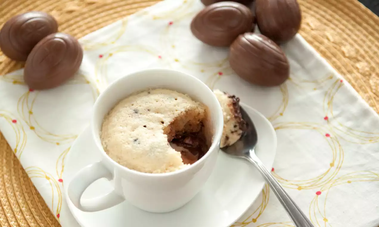 Lichtgele mug cake met luchtige textuur en gesmolten chocoladevulling in een witte mok met een lepel op een bord, omringd door chocolade-eitjes