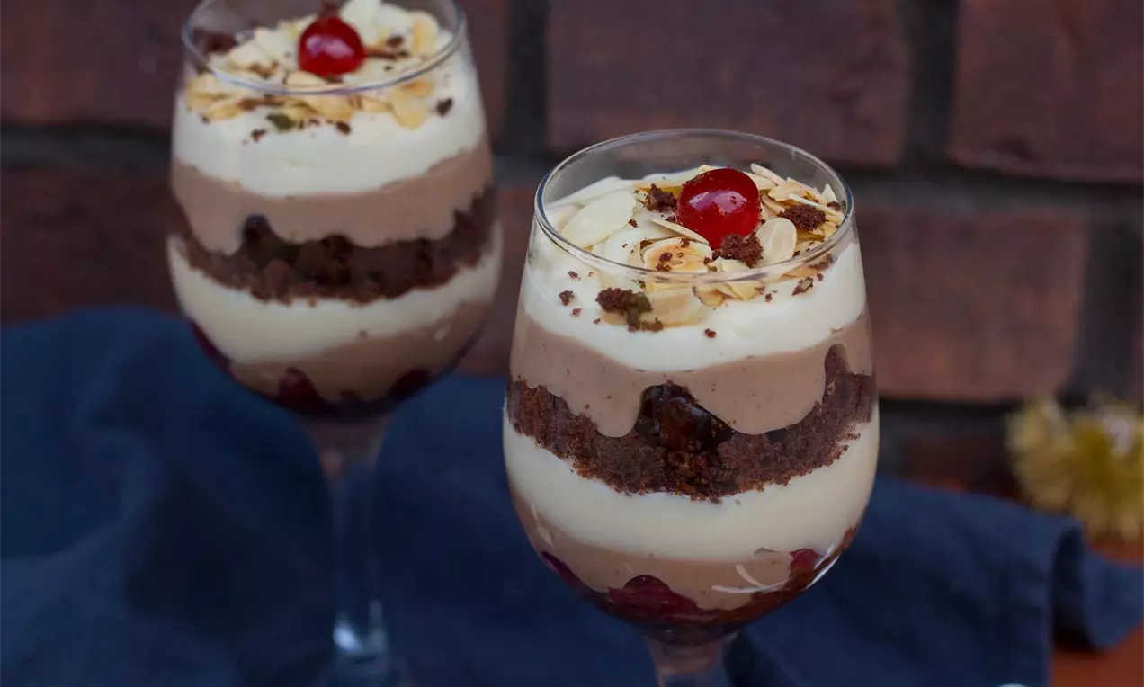Vysoké sklenice vrstvené světlým krémem, kaštanovým mousse, tmavým korpusem, šlehačkou, plátky mandlí a červenou třešní na vrcholu