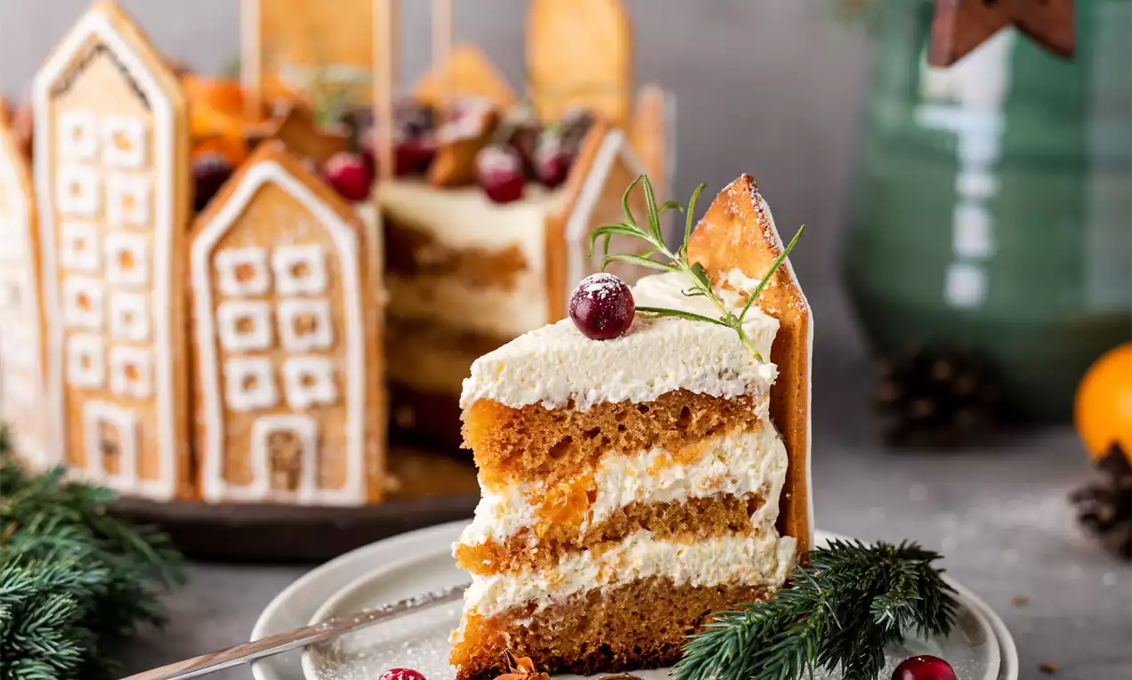 Trijų sluoksnių medaus torto gabalėlis su šviesia kremine tekstūra, papuoštas rozmarino šakele ir spanguolėmis ant pilkos lėkštės, šalia eglių šakelių