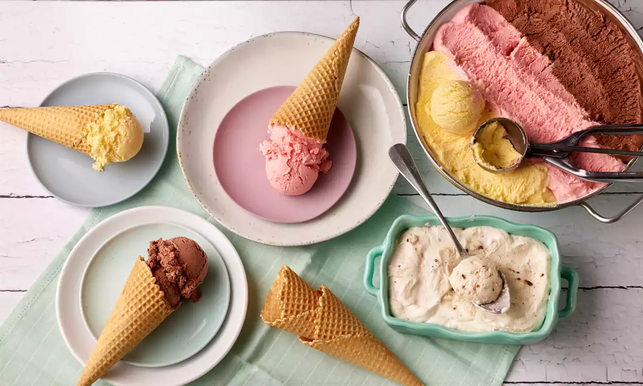 Eiswaffeln mit Kugeln in Rosa, Gelb, Weiß und Braun, angerichtet auf verschiedenen Tellern, daneben Schalen mit Eis und Eiskugelzange