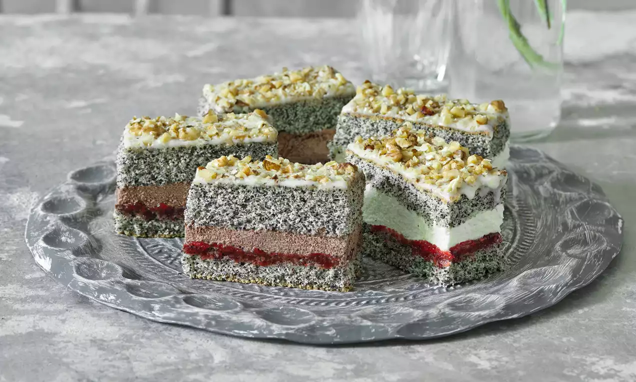 Čtvercové řezy s šedým makovým těstem, vrstvy červené marmelády a světlého krému, posypané drcenými ořechy na stříbrném talíři