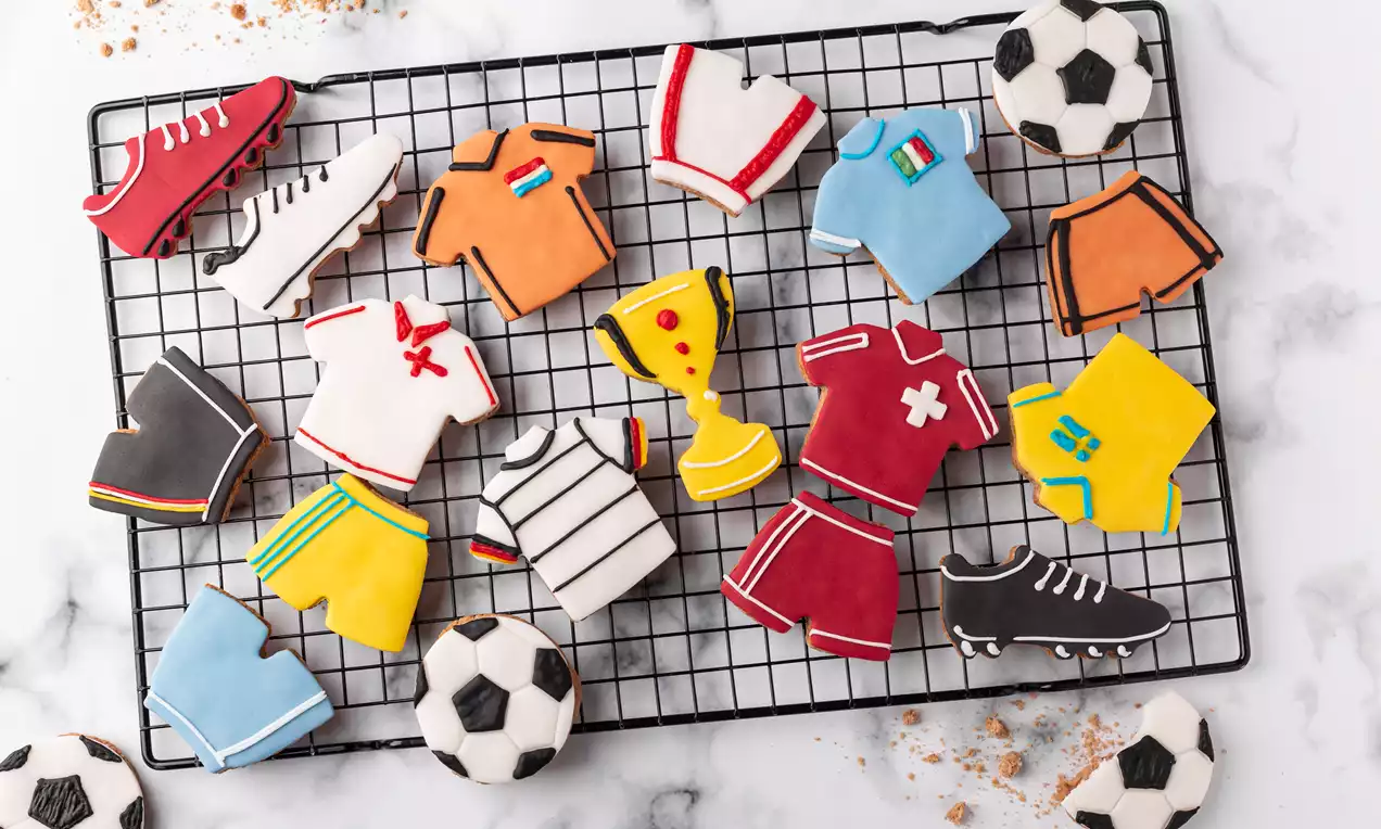 Süsse Guetzli in Trikot- und Fußballformen bunt verziert auf einem schwarzen Kuchengitter mit Krümeln und einzelnen Fußballkeksen auf Mamor