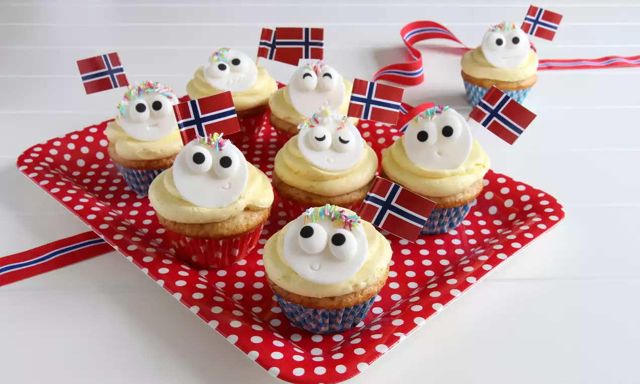 Små muffins i fargerike former med lys gulfarget glasur, øyne av sukkermasse, strøssel og norske flagg på rødt, prikkete brett