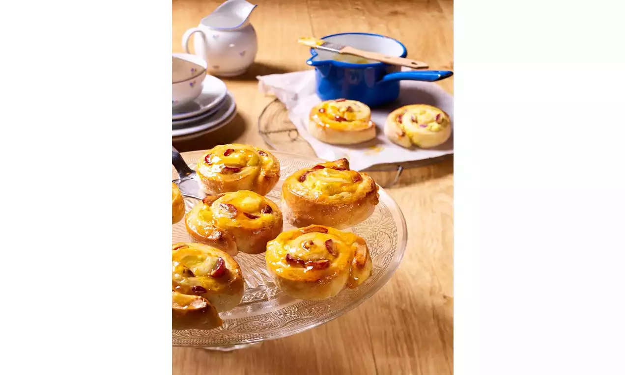 Goldgelbe Traubenschnecken mit glänzender Oberfläche und leichter Bräune auf einer gläsernen Tortenplatte, im Hintergrund Geschirr und ein blauer Topf