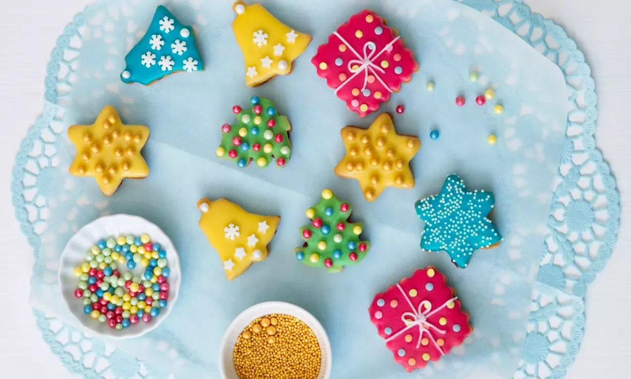 Farebné vianočné sušienky v tvare stromčeka, hviezdy, zvončeka a darčeka, zdobené cukrovou polevou a posypom na modrej servítke