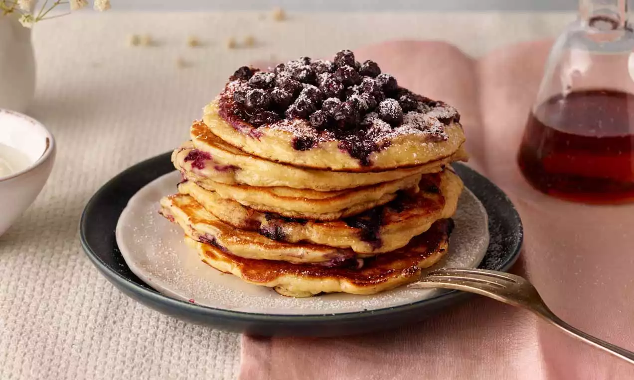 Ein Stapel goldbrauner Pancakes mit saftigen Heidelbeeren und Puderzucker auf einem dunklen Teller, daneben Ahornsirup und ein rosa Stoffserviette