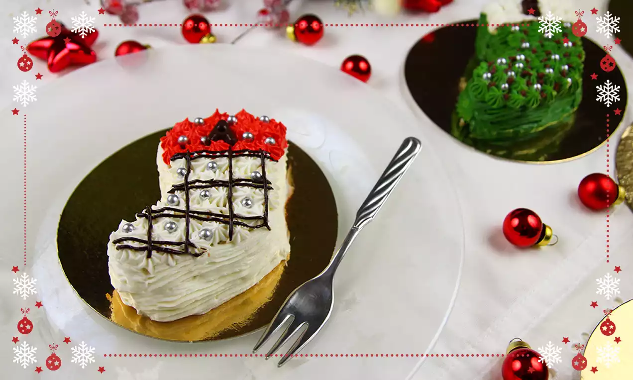 Tortina a forma di cuore con base dorata, crema bianca ricamata, dettagli rossi e argento, adagiata su piattino trasparente con forchettina