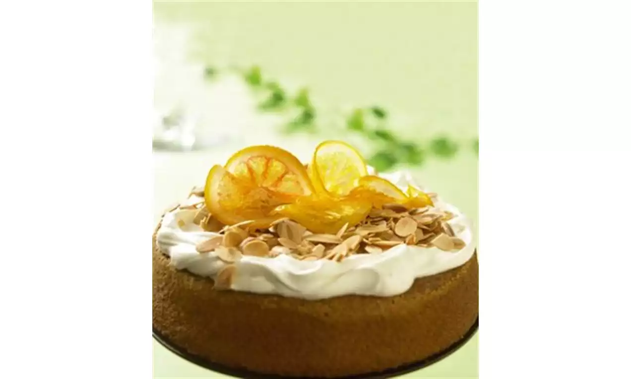 Mandel- og appelsinkake med gyllen brun kant, hvit kremtopping, ristede mandelflak og skiver av kandisert appelsin på grønn bakgrunn