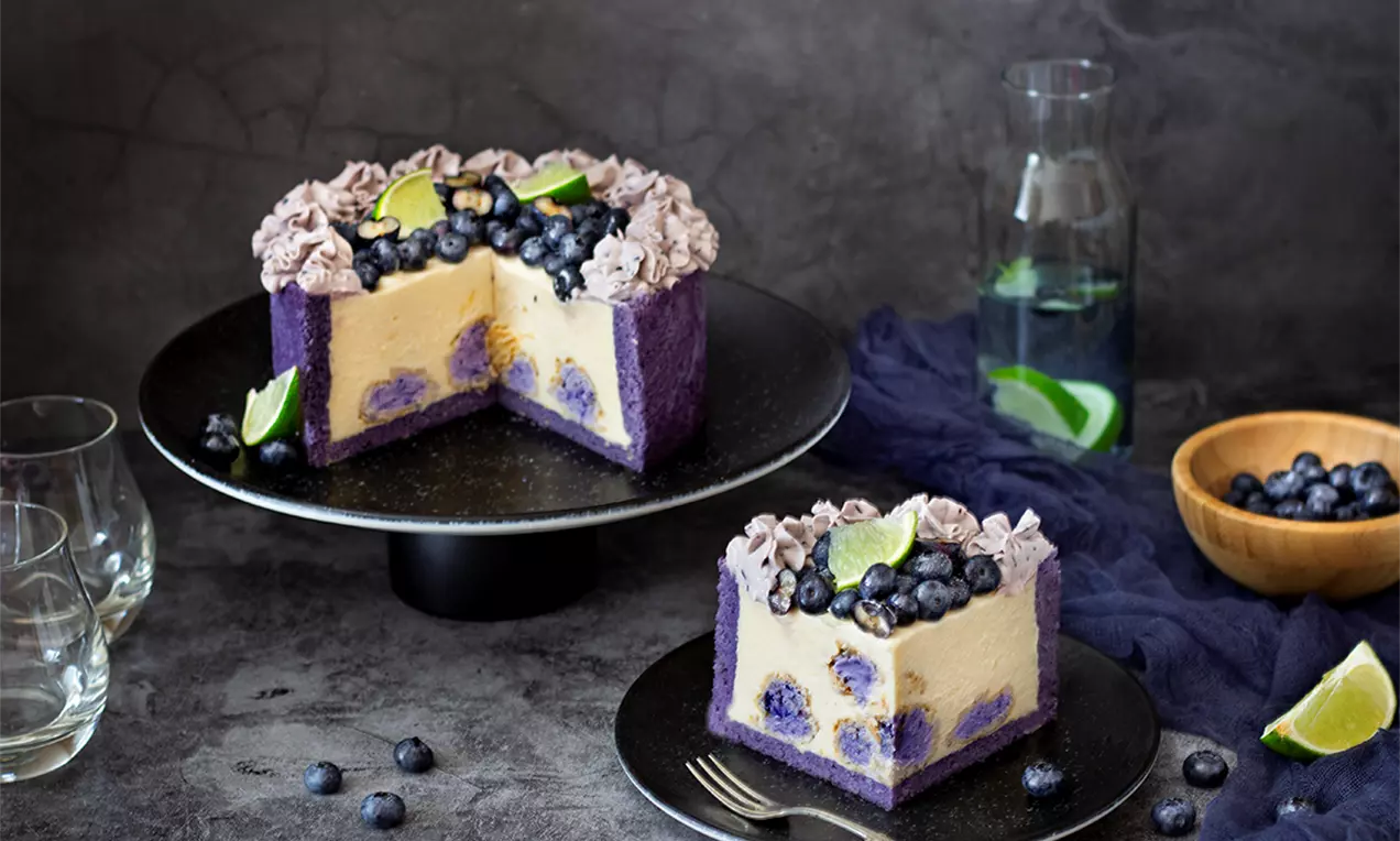 Боровинкова торта с профитероли, с лилав блат и светъл крем, украсена със свежи боровинки, лайм и разбита сметана върху тъмен поднос