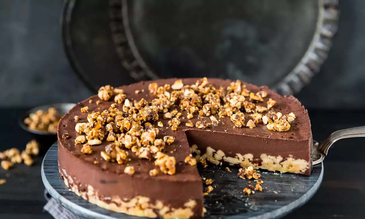 Čokoladna torta temno rjave barve z gladko površino, obložena z zlatimi hrustljavimi lešniki na steklenem podstavku s sivim prtičem