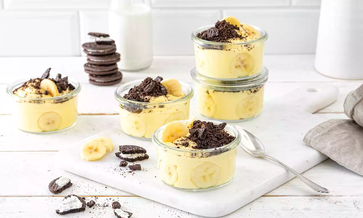 Dessert im Glas mit gelber Bananencreme, frischen Bananenscheiben und dunklen Keksbröseln, angerichtet auf weißem Untergrund mit Löffel