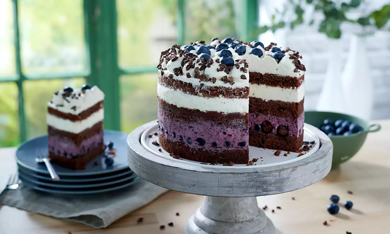 Troslojna torta sa čokoladnim biskvitom, kremom od jogurta i borovnica, ukrašena svježim borovnicama i čokoladnim listićima na bijelom stalku