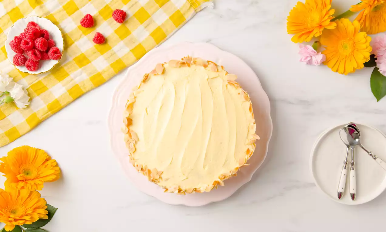 Tarta de crema amarilla pálida con bordes irregulares dorados sobre plato rosa, rodeada de flores y frambuesas junto a cubiertos sobre mantel amarillo