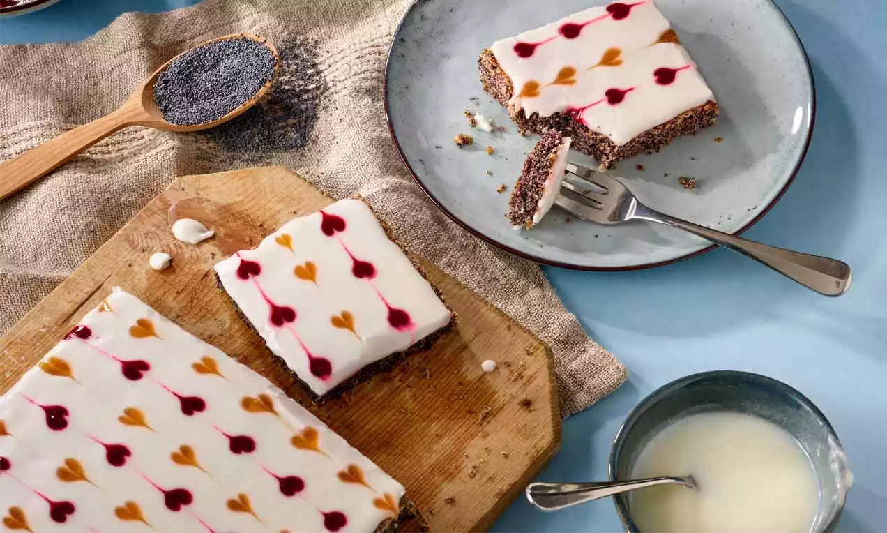 Mohnkuchen mit weißer Glasur und bunten Herzmustern, rechteckig geschnitten, auf Holzbrett und grauem Teller mit Mohn und Glasur angerichtet
