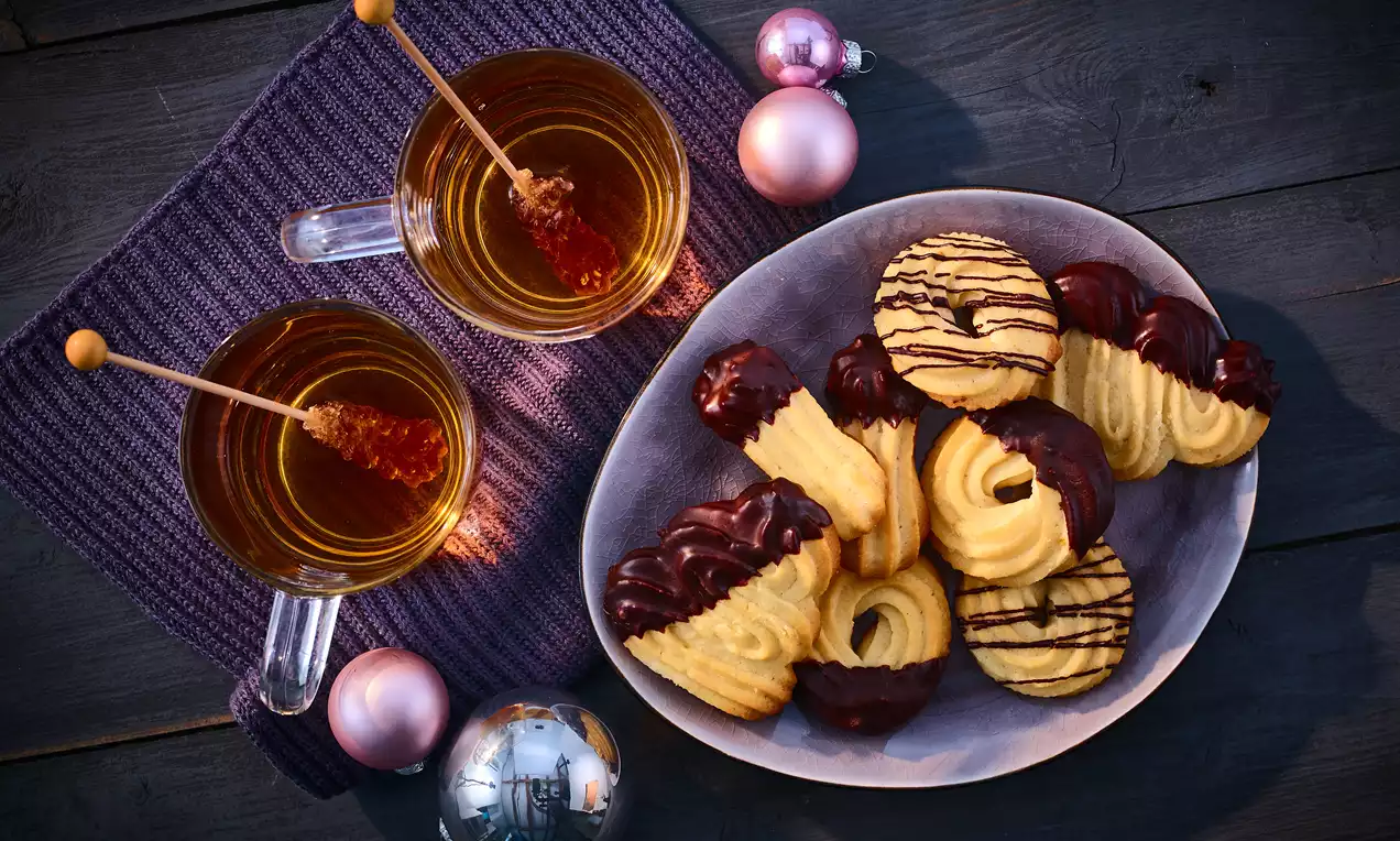 Veganes Spritzgebäck in geschwungenen Formen mit dunkler Schokolade verziert auf grauem Teller, daneben Tee in Gläsern mit Kandissticks