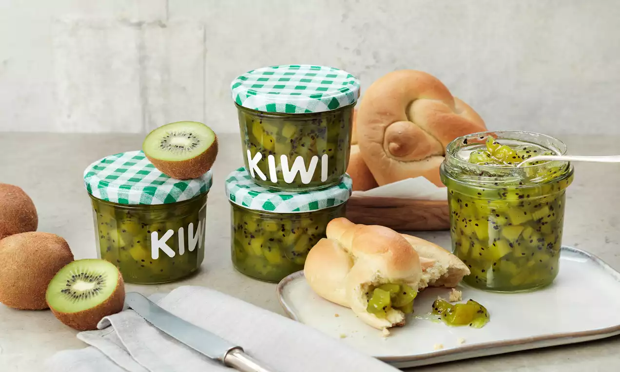 Gläser mit grün-gelber Kiwimarmelade und Deckeln im Karomuster, daneben aufgeklapptes Brötchen und frische Kiwis auf grauem Untergrund