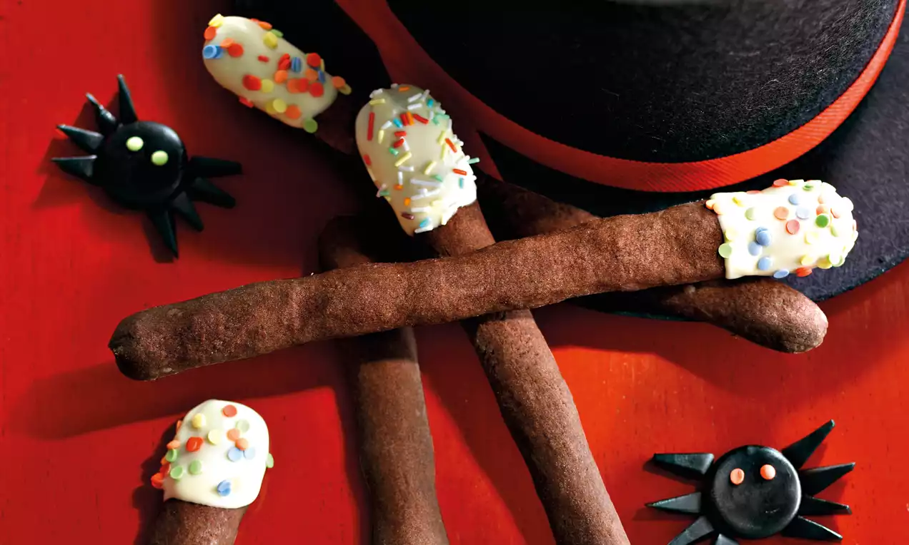Schokoladenbraune Stäbe mit weißer Glasur und bunten Streuseln an den Enden, arrangiert auf rotem Hintergrund mit Hut und schwarzer Dekospinne