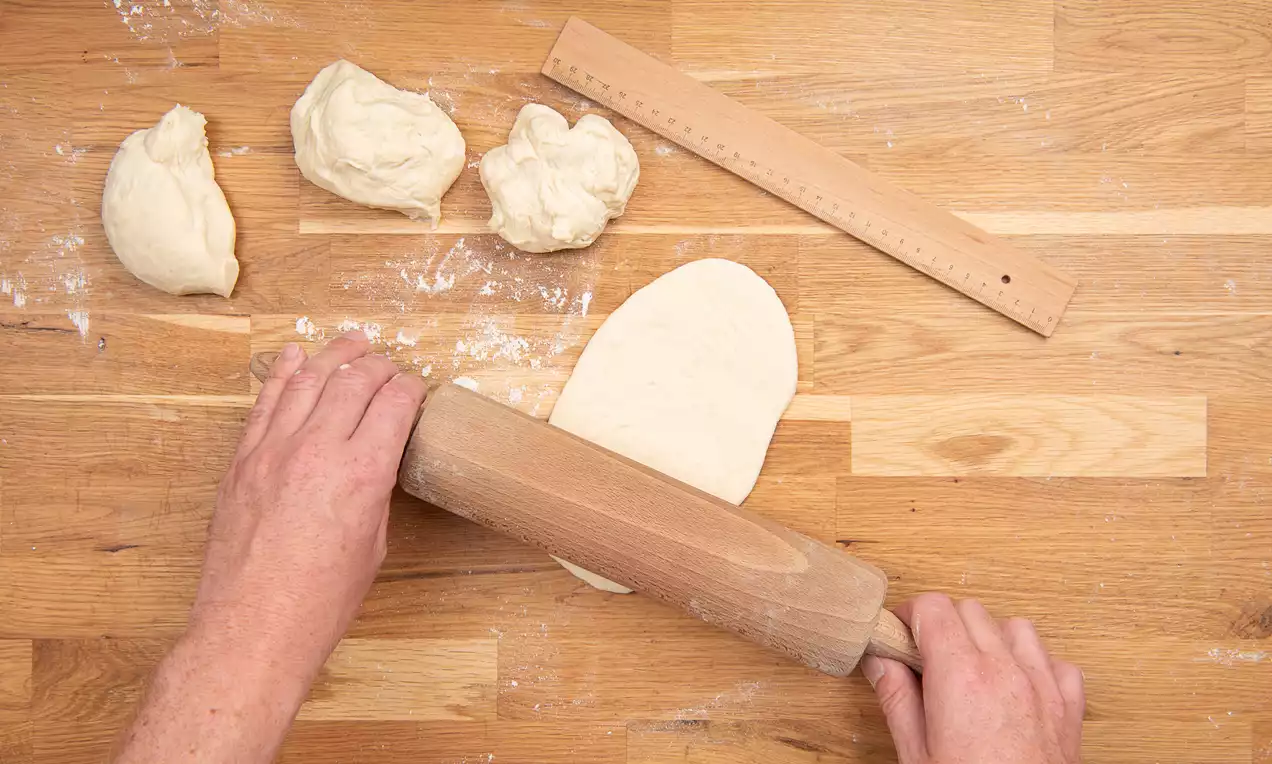 Deux mains étalent une pâte beige souple avec un rouleau en bois sur une surface en bois clair, entourée de morceaux de pâte farinés et d'une règle.