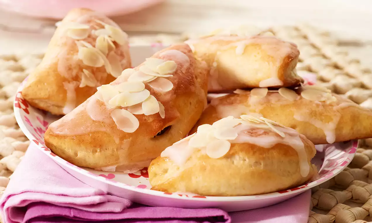 Chaussons dorés en forme de triangles, garnis de glaçage brillant et d’amandes effilées, servis sur une assiette fleurie rose