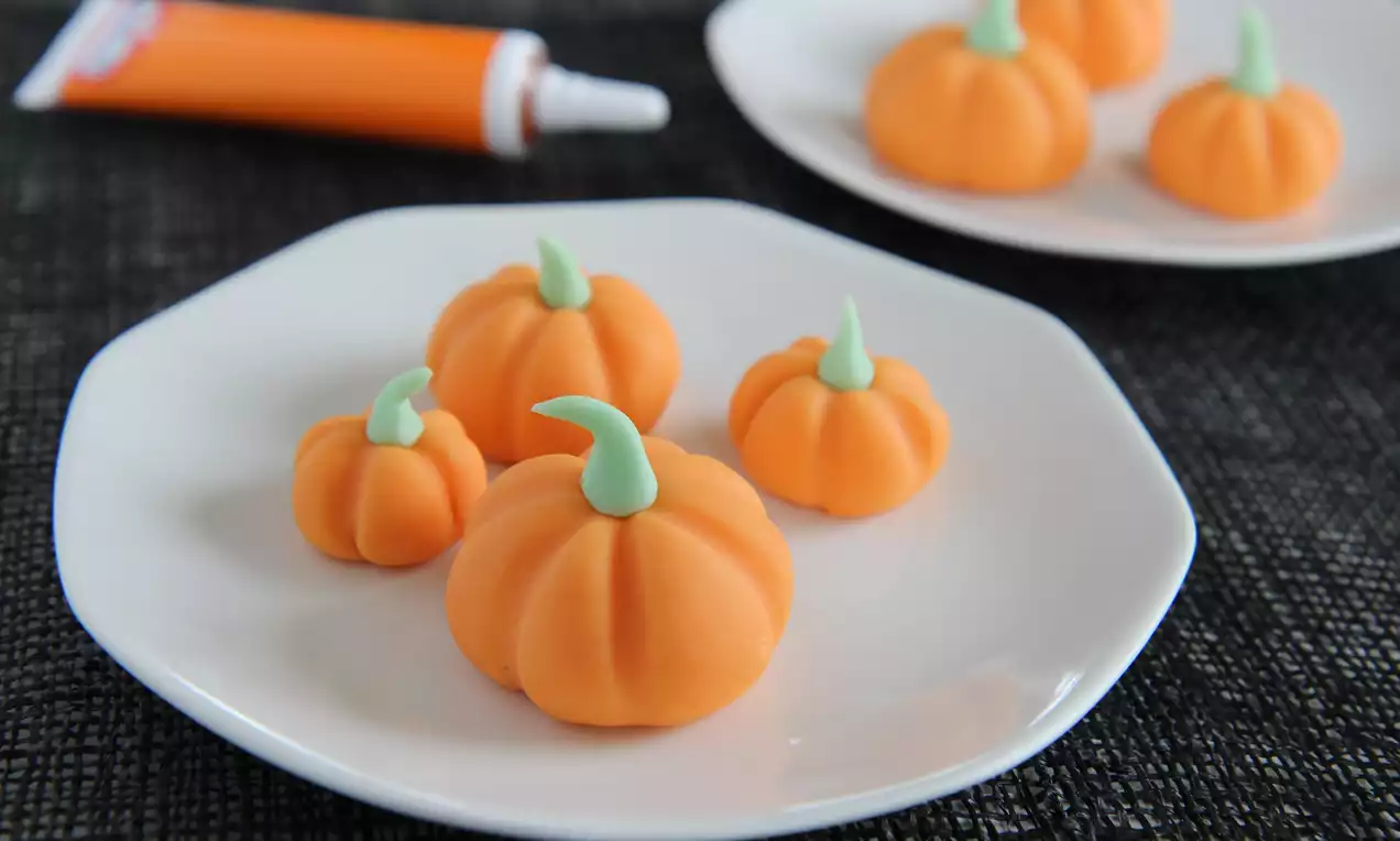 Små gresskar av oransje fondant med lysegrønne stilker, plassert på hvite tallerkener mot mørk teksturert bakgrunn