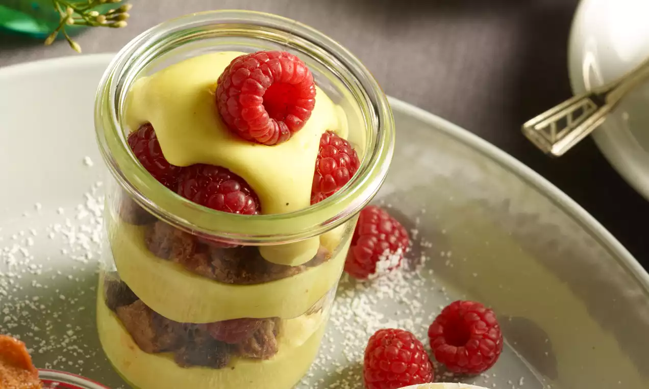 Pudding trifle in een glazen potje met lagen van gele pudding, chocoladecakeblokjes en helderrode frambozen, geserveerd op een licht bord