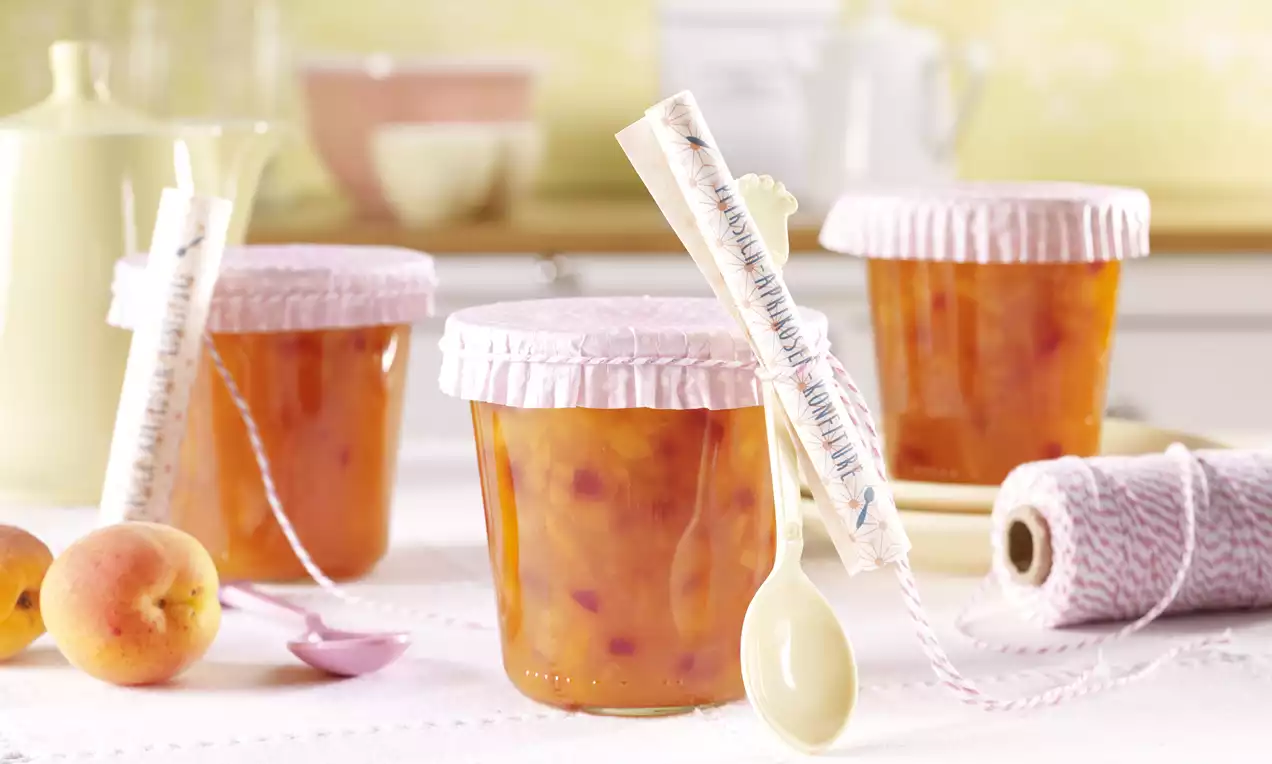 Staklenke punjene zlatno-narančastom marmeladom s tamnocrvenim komadićima voća, poklopljene ružičastim papirnatim omotom i ukrašene žlicama