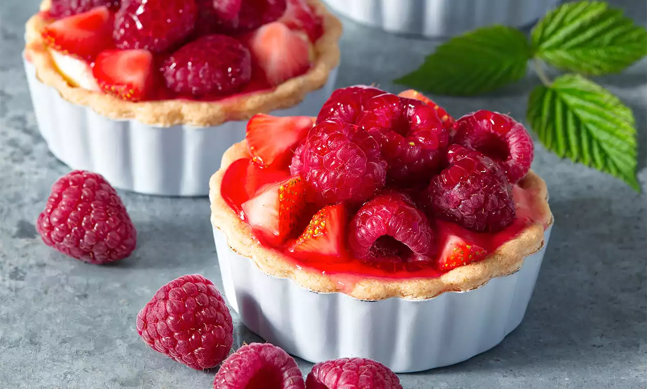 Bezlepkové košíčky s jemnou světlou křupavou krustou, plněné krémem a čerstvým ovocem malin a jahod na šedém podkladu s lístky